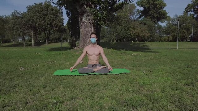 Beautiful fit young man with mask practice yoga in the park, brunette man doing exercise and spending active time