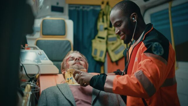 Happy Black African American Paramedic Poses for Camera and Gives a Thumbs Up in an Ambulance Vehicle with an Injured Male Patient. Emergency Medical Technician is Using a Stethoscope.