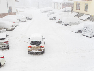 The car drives through a snow-covered yard in a blizzard past parked cars covered with snow