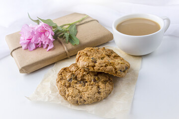 Romantic vintage still life with pretty gift box wrapped with brown craft paper and decorated with pink flower on white background. Dessert (oat cookies) served for tea or coffee break.