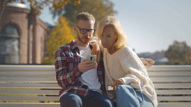 Young man showing photo on cellphone to mature mother relaxing on city bench