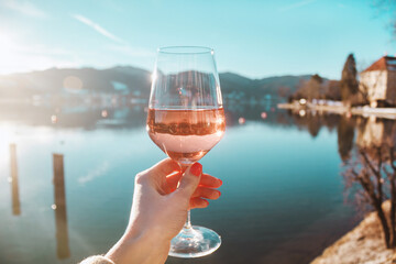 Female hand with glass of rose wine. Cozy pier on the coast of the lake Tegernsee. Alps mountains...