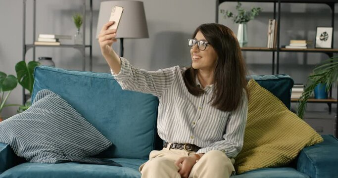 Pretty young Caucasian woman in glasses sitting on couch in room, smiling and taking selfie photo on smartphone. Beautiful happy smiled girl making photos with phone camera. Female smiling and posing.