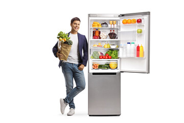Full length portrait of a young man with a grocery bag leaning on an open fridge filled with food
