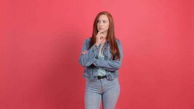 Serious young woman sits with crossed hands and looks aside as she thinks about important things, isolated over red background.