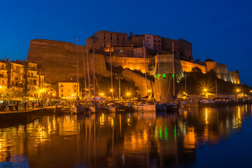Fototapeta na wymiar Hafen mit Schiffen (Motor- und Segelboote) und die Zitadelle mit blauem Himmel in Calvi, Hauptort der Balagne, auf der französischen Mittelmeerinsel Korsika. Nachtaufnahme mit Beleuchtung.