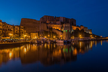Hafen mit Schiffen (Motor- und Segelboote) und die Zitadelle mit blauem Himmel in Calvi, Hauptort der Balagne, auf der französischen Mittelmeerinsel Korsika. Nachtaufnahme mit Beleuchtung.