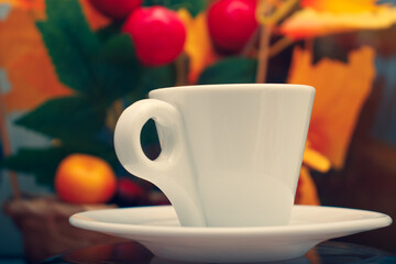 White porcelain conical cup with an eyelet on a saucer close-up. A cup on the background of a basket with a bouquet of imitation of branches of autumn trees with yellowed leaves.