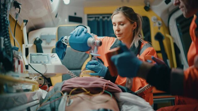EMS Paramedics Team Provide Medical Help to Injured Patient on the Way to Hospital. Emergency Care Assistants Using a Defibrillator and Ventilation Mask to Bring the Man Back to Life in an Ambulance.