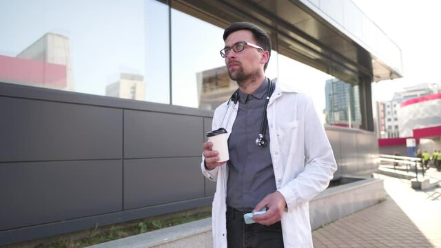 Medical professional during coffee break after long day. General practitioner drinking coffee to go while walking on the background of the clinic. Doctor in a lab coat drinks a hot drink