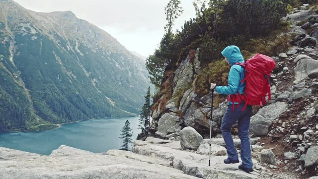 Hiker woman walking with backpack admiring mountain lake view landscape from the cliff edge. Stabilized, 4K Ultra HD. Epic Steadicam hiking in a stormy wind, lifestyle. Misty Mountains Series. 