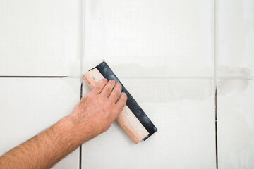 Worker hand using rubber trowel and grouting seams with paste between gray ceramic tiles on floor....