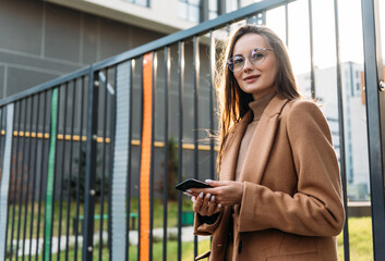 Fototapeta premium Smiling curly businesswoman wearing trendy sunglasses walks down the central city street and uses her phone. Businesswoman holding cell phone.