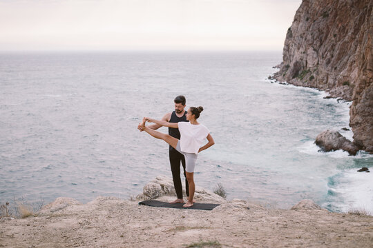 Young man coach teaches woman yoga exercises on an outdoors class on mountain with beautiful sea view.