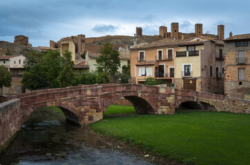 Fototapeta premium Scenic view of a medieval bridge made with reddish stone blocks and the castle on the top of the hill behind the houses on a cloudy day, Molina de Aragón, Guadalajara, Spain