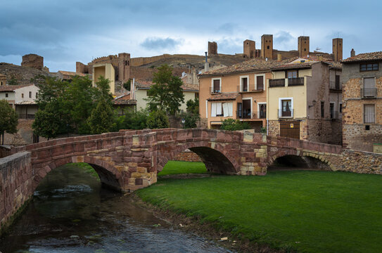 Scenic view of a medieval bridge made with reddish stone blocks and the castle on the top of the hill behind the houses on a cloudy day, Molina de Aragón, Guadalajara, Spain