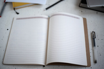 An open notebook with blank pages and a black pen on a light background. Diary, book, and tablet in the background. Top view. Space for text. Flat lay workspace and planning.