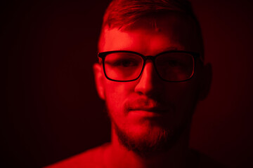 Low key portrait of bearded young man in glasses full-face looking at camera in red light soft focus on black backdrop. Loneliness, sleeplessness concept. Night torments of creativity