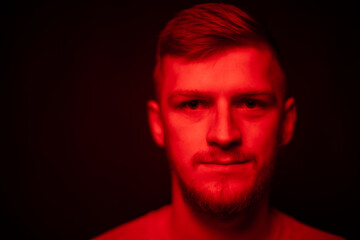 Low key portrait of bearded young man full-face looking at camera dilated pupil in red light on...