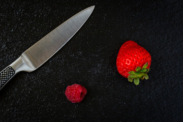 A strawberry and a raspberry next to kitchen knife placed on embossed black stone background