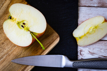 Half of an apple placed on cutting board over black stone and a quarter of an apple on wooden table with a stainless still knife intersecting all those surfaces.
