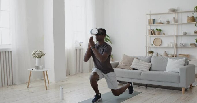 African american guy in VR headset doing lunges forward exercise at home, watching online video tutorial - Powered by Adobe