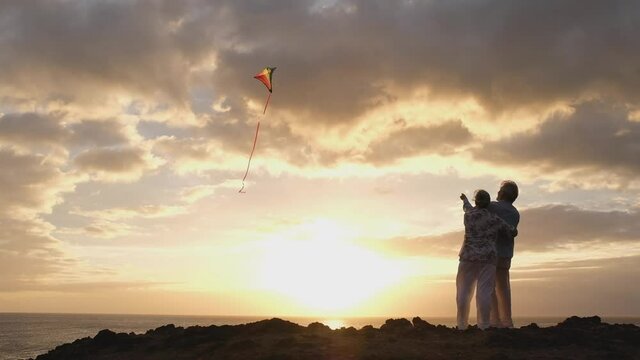 close up and portrait of two old and mature people playing and enjoying with a flaying kite at the beach with the sea at the background with sunset - active seniors having fun
