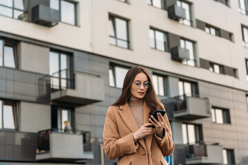 Fototapeta premium Smiling curly businesswoman wearing trendy sunglasses walks down the central city street and uses her phone. Businesswoman holding cell phone.
