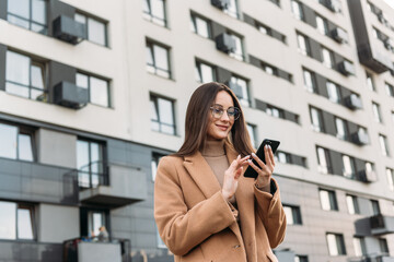 Happy pretty brunette girl using mobile phone near office, beautiful woman browsing phone and doing online shopping in a mobile app while smiling walking near shopping mall.