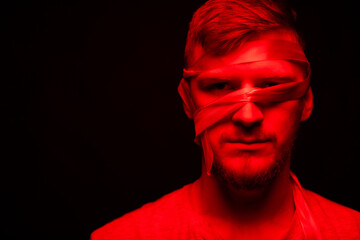 Low key portrait of bearded young bandaged face man full-face looking at camera in red light on black backdrop. personal expression Restrictions, social isolation, segregation and dissociation concept