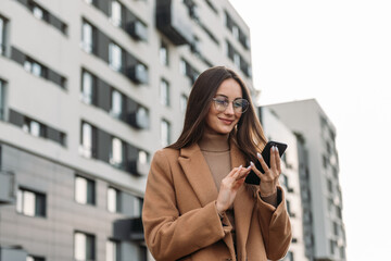 Smiling curly businesswoman wearing trendy sunglasses walks down the central city street and uses her phone. Businesswoman holding cell phone.