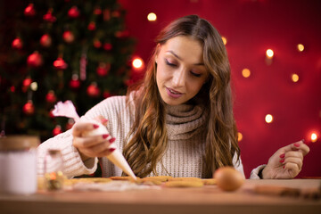 Christmas baking. pretty brunette young woman icing Christmas gingerbread on red studio background with Christmas tree and Xmas decoration. Festive food, family culinary concept