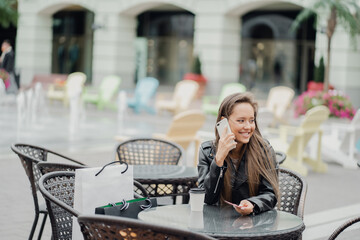 Fototapeta premium a confident, European-looking brunette woman in a black leather jacket and blue jeans looks away. sitting on a bench next to bags sale clothes in the outlet shopping buy