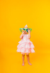 a cheerful little girl in a pink puffy dress holds big funny glasses in her hands and blows a whistle on a yellow background