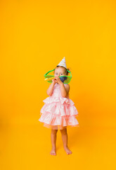 A curious little girl in a festive dress and cap stands in large glasses on a yellow background with a copy of space