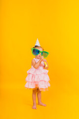 a full-length little girl stands in a paper hat with big funny glasses and blows a yellow whistle on a yellow background with space for text