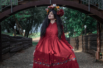 beautiful young girl in a red dress with long sleeves and a wreath is dancing under the bridge in the park
