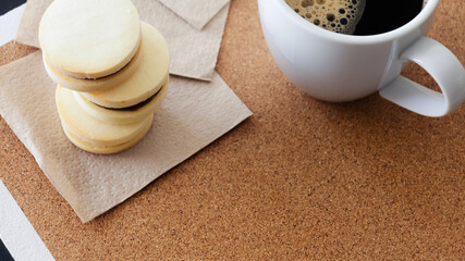 Primer plano desde un &aacute;ngulo cenital de una taza de cer&aacute;mica blanca de caf&eacute; negro caliente junto a tres peque&ntilde;os alfajores rellenos de dulce leche horneados en casa y bajo iluminaci&oacute;n natural. 