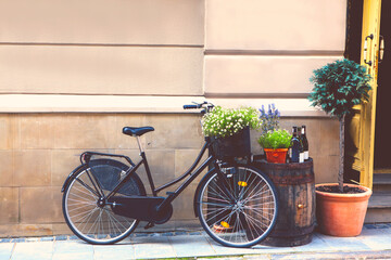Black retro bicycle stands next to the barrel, flower pots, wine bottles, on the street, next to the open vintage door, against the background of a stone wall, with a free space for text.