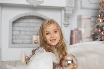Little cute girl with blond hair with angel wings in the studio next to the Christmas tree and with Christmas balls
