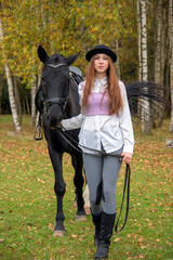 Red-haired girl in a black hat with a horse in the autumn forest.