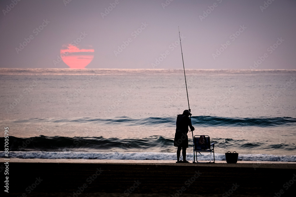 Wall mural man fishing in the sea with the sun in the background