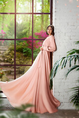 slender young woman in a beautiful pink long dress  stands at the big window