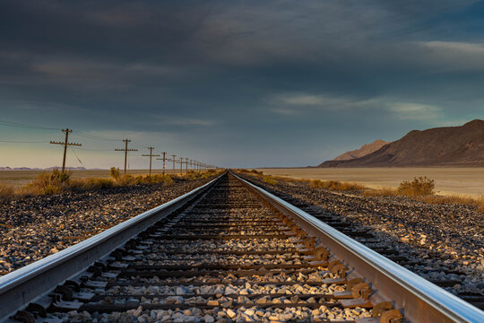 A nice landscape image of an old railroad track in the desert with a telegraph line next to it and mountains in the background. This beautiful image was taken during a gold hour sunset.