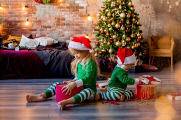 Two young children in pajamas and Santa hats unpack Christmas presents in the room