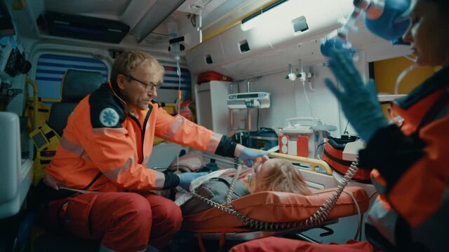 EMS Paramedics Team Provide Medical Help to Injured Patient on the Way to Hospital. Emergency Care Assistants Using a Defibrillator and Ventilation Mask to Bring the Man Back to Life in an Ambulance.