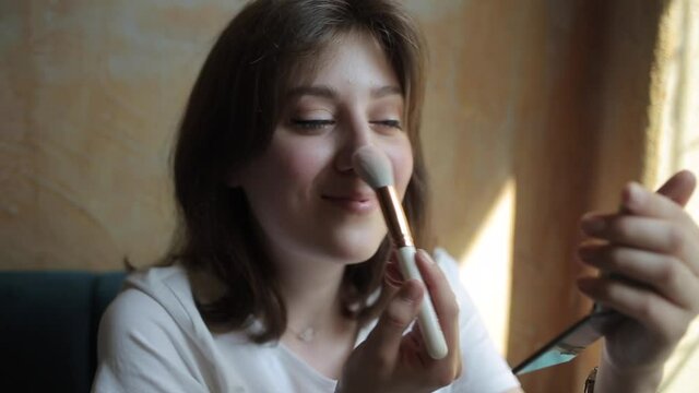 Cheerful young woman in a white T-shirt does makeup and applies powder to her face with a brush while looking in the mirror. Front view. Close-up. Camera zooms in and out