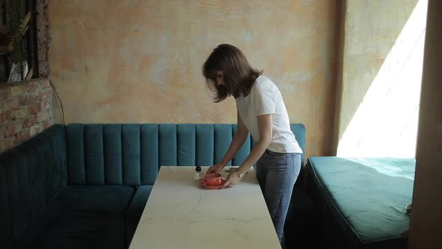 A beautiful young girl in a white T-shirt and jeans makes a layout for flat lay photos on a mobile phone from cosmetics and a cup while standing at a cafe table. Side view