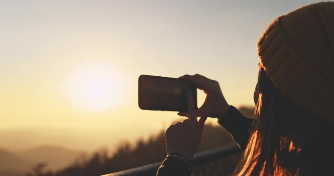 Young Woman Taking Pictures of Sunlit Fall Mountain Landscape form the Top. SLOW MOTION. Hiker Girl is Taking photos with smartphone of autumn hills landscape at sunset, Lens Flare.  