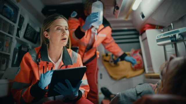 Female EMS Professional Paramedic Using Tablet Computer to Fill a Questionnaire for the Injured Patient on the Way to Hospital. Emergency Care Assistant Comforting the Patient in an Ambulance.